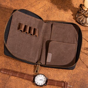 May include: Open, brown leather travel case with a black zipper, featuring multiple pen holders and a small pocket. A brown leather watch with a white face and Roman numerals is next to the case. The watch band has a crocodile pattern.