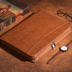 May include: A brown leather zipped portfolio with a silver zipper pull. The portfolio is lying on a brown wooden surface with a book, glasses, and a watch.