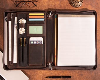 May include: Open brown leather portfolio with multiple compartments, including card slots, a phone pocket, and a notepad. The portfolio is open, revealing a white notepad, a smartphone, and a laptop. A pair of glasses and a candle holder are on the table.