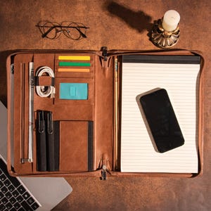 May include: A brown leather portfolio with a zipper closure, multiple pockets, and a notepad. The portfolio is open and has a phone, a pen, and a card holder inside.