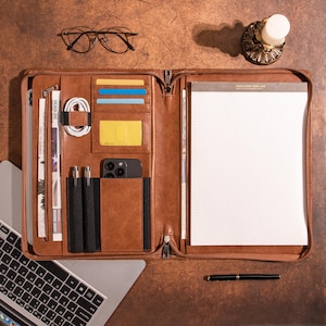 May include: A brown leather zippered portfolio with multiple pockets and compartments. The portfolio is open and contains a notepad, pens, a phone, and cards. The portfolio is on a brown wooden surface.