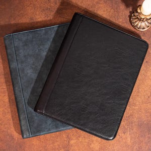 Könnte beinhalten: Zwei Dokumentenmappen aus Leder. Eine ist dunkelgrau-blau mit hellblauen Nähten, die andere schwarz mit einem braunen Rücken. Die Mappen sind rechteckig und liegen auf einer braunen Oberfläche.