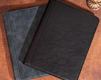 May include: Two leather document folders. One is a dark gray-blue with light blue stitching, and the other is black with a brown spine. The folders are rectangular and lie on a brown surface.