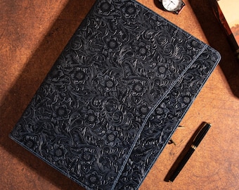 May include: A dark blue leather portfolio with an embossed floral pattern. The portfolio is closed and has a clasp. A brown leather watch and a black pen with gold accents are also visible.