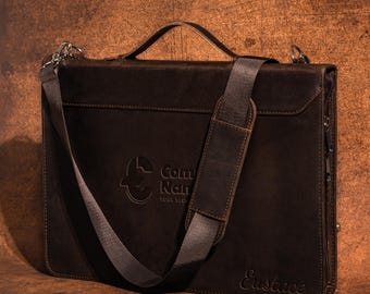 May include: A dark brown leather briefcase featuring a top handle and an adjustable shoulder strap. The front displays an embossed circular logo with the text Company Name and the word Eustace in a cursive script font at the bottom right corner.