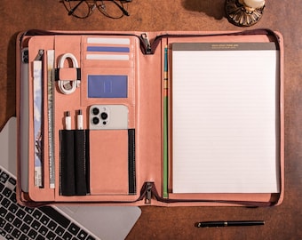 May include: A pink leather portfolio with multiple compartments, including card slots, a phone pocket, and a notepad. The portfolio is open, revealing a notepad, a phone, and a charging cable. A laptop and glasses are also visible.