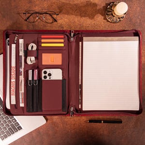 Può includere: Una cartella portadocumenti con cerniera in similpelle bordeaux, dotata di scomparti interni per carte, uno smartphone, penne e un cavo. Il lato sinistro presenta una tasca verticale con cerniera recante la scritta MAGNIS DISARE, mentre il lato destro contiene un blocco note a righe.