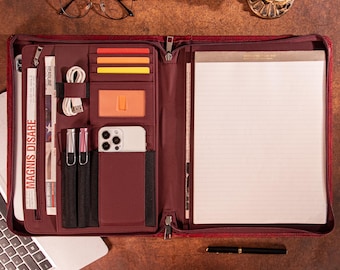 May include: A burgundy leather-textured zippered portfolio organizer featuring multiple interior slots for cards, a smartphone, pens, and a cable. The left side includes a vertical zippered pocket with the text MAGNIS DISARE, while the right side holds a lined notepad.