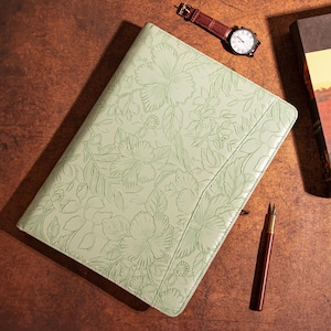 May include: A light green leather bound journal with a floral embossed design. The journal is closed and lying flat on a brown surface.