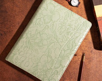 May include: A light green leather journal with embossed floral design. A brown leather watch and a fountain pen are next to the journal. The journal is on a brown wooden surface.