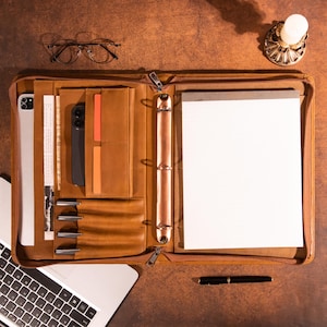 Könnte beinhalten: Eine braune Ledermappe mit mehreren Fächern, darunter eine Tablettasche, Stifthalter und ein Notizblock. Die Mappe ist geöffnet und zeigt ein Smartphone, Stifte und einen leeren Notizblock.