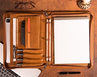 May include: A brown leather portfolio with multiple compartments, including a tablet sleeve, pen holders, and a notepad. The portfolio is open, revealing a smartphone, pens, and a blank notepad. A laptop and glasses are also visible.