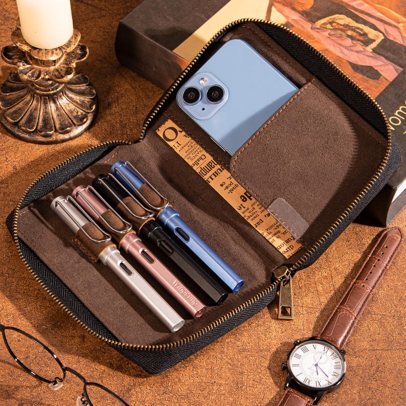 May include: A brown leather zippered case with a blue smartphone, several pens, and a brown leather watch. The pens are silver, pink, black, and blue. The watch has a white face and Roman numerals.