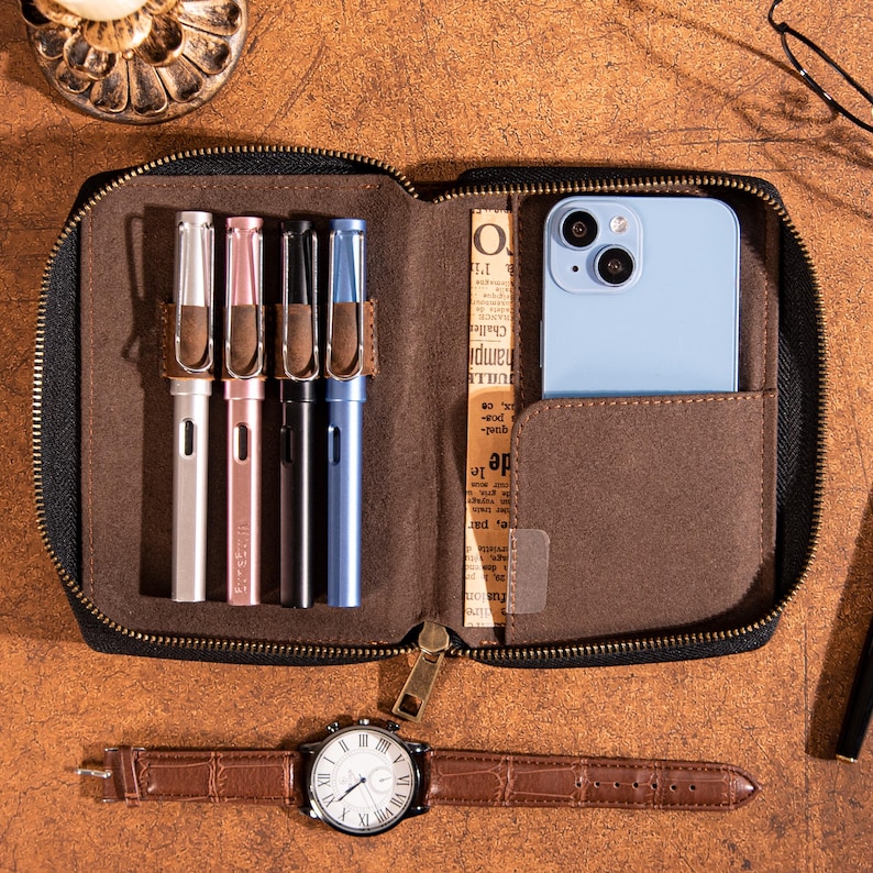 May include: A brown leather pen case with a zipper, containing four pens in silver, pink, black, and blue. A light blue smartphone is in a pocket. A brown leather watch with a silver face and brown strap is below.