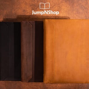 Può includere: Tre custodie per laptop in pelle di diversi colori: nero, marrone e beige. Le custodie sono allineate una accanto all'altra su una superficie marrone testurizzata. Il testo "JumpNShop" è visibile sopra le custodie.