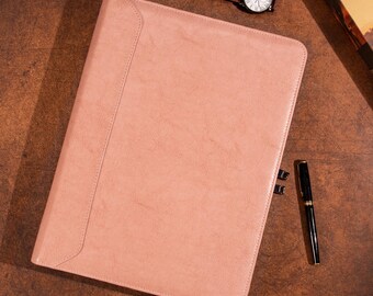 May include: A blush-colored leather portfolio with a flap closure. A brown leather watch with a white face and a black fountain pen are next to the portfolio. The items are on a brown wooden surface.