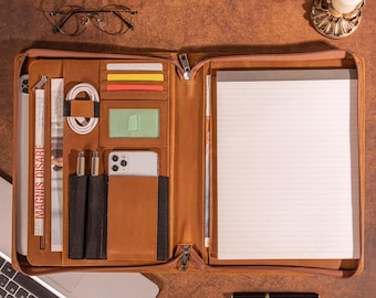 May include: A brown leather portfolio with multiple compartments, including card slots, pen holders, and a phone pocket. It contains a notepad, a smartphone, and a laptop. The portfolio is open, revealing its contents, and is zipped halfway.