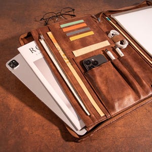 May include: A brown leather portfolio with multiple pockets and compartments. The portfolio is open and contains a tablet, a phone, a magazine, cards, and pens.