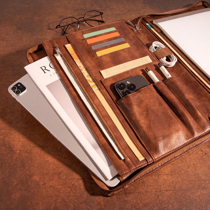 May include: A brown leather portfolio with multiple pockets and compartments. The portfolio is open and contains a tablet, a phone, a magazine, cards, and pens.