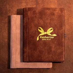May include: A dark brown rectangular portfolio with a gold-coloured ribbon graphic, the name Katherine, and the date 2023.10.17 printed on the front cover, layered over a light pink portfolio of the same rectangular shape and design.