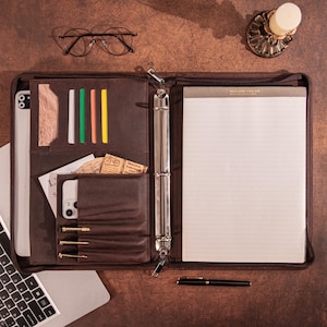 May include: A brown leather portfolio with a zipper, open to reveal a notepad, pen, and various pockets holding a phone, cards, and a wooden accent. A laptop and glasses are also visible. The overall theme is business and organization.