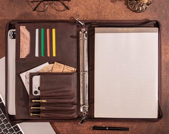 May include: A brown leather portfolio with a zipper, open to reveal a notepad, pen, and various pockets holding a phone, cards, and a wooden accent. A laptop and glasses are also visible. The overall theme is business and organization.