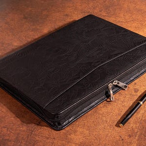 May include: A black leather zipped portfolio with an embossed floral design. The portfolio is lying on a brown surface with a gold pen next to it.