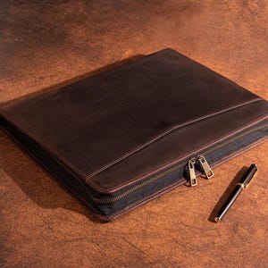 May include: A dark brown leather portfolio with a black zipper and two gold-colored zipper pulls. A black and gold pen rests beside the portfolio. The portfolio is rectangular and appears to be closed.