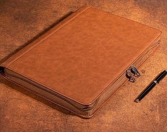 May include: A brown leather portfolio with a zipper closure and a black pen with gold accents. The portfolio has a textured surface and is rectangular in shape. The pen is lying on a brown surface.