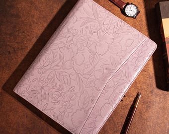 May include: A light pink leather portfolio with an embossed floral design. A brown leather watch with a white face and a brown pen are next to the portfolio. A book is in the background.
