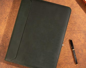 May include: Dark green leather portfolio with a flap closure, lying on a brown surface. A brown leather wristwatch and a black pen with gold accents are also visible. The portfolio is a business accessory.