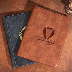 May include: A brown leather-like portfolio featuring a tuxedo graphic and the name Charlotte engraved on the front, layered over a dark gray portfolio with a yellow laurel wreath design. Both items have visible stitching along the edges.