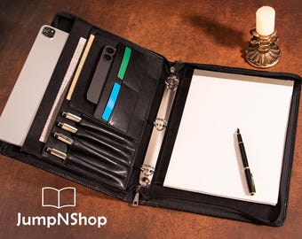 May include: Black leather portfolio with a zipper closure, open to reveal a tablet, pens, notepad, and card slots. A pen rests on the notepad. The JumpNShop logo is visible at the bottom.