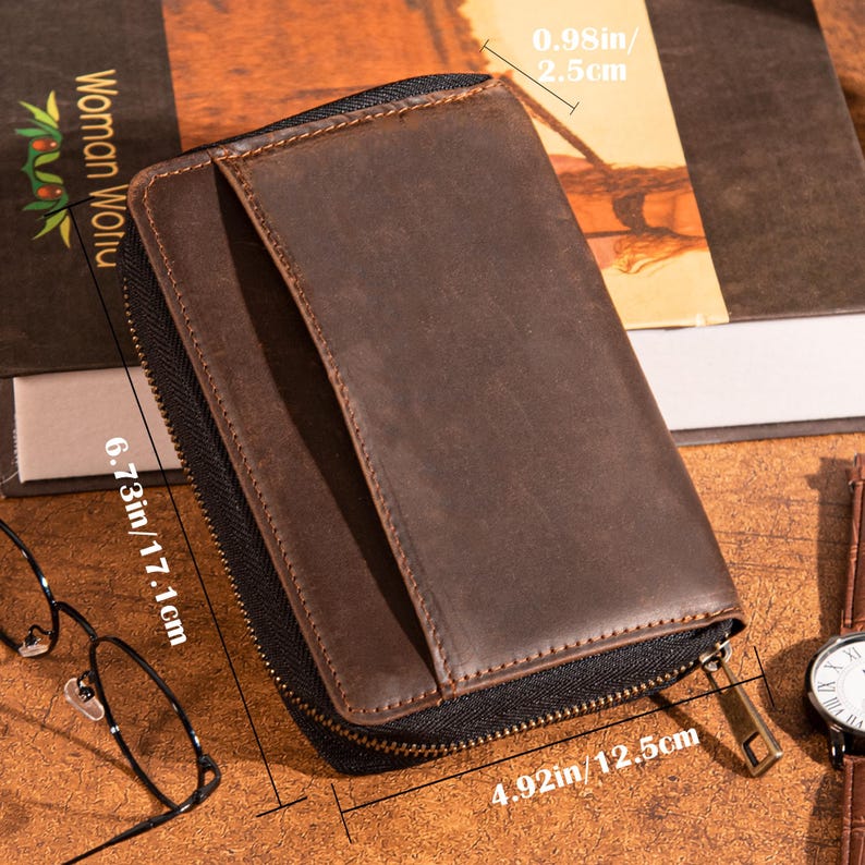 May include: A brown leather wallet with a zipper closure. The wallet measures 6.73 inches (17.1 cm) in height, 4.92 inches (12.5 cm) in width, and 0.98 inches (2.5 cm) in thickness. The wallet is on a wooden surface.