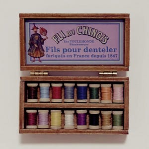 Può includere: Un'antica scatola da cucito in legno con coperchio a cerniera, con un'etichetta con testo francese e un'illustrazione. La scatola contiene due file di colorate bobine di filo. Il testo sull'etichetta recita "Fils au Chinois" e "fabriqués en France depuis 1847".