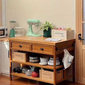 Może przedstawiać: Drewniana wyspa kuchenna z dwoma szufladami i dwoma półkami. Wyspa ma jasne wykończenie drewna i czarne metalowe okucia. Na wyspie znajduje się zielony mikser stojący, blender, drewniana skrzynia z etykietą "WOODENBOX NO.1" oraz różne naczynia na półkach.