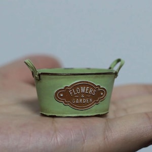 May include: A small, light green metal planter with two handles. The planter has a brown label that reads "FLOWERS & GARDEN". The planter is designed to hold small plants or flowers.