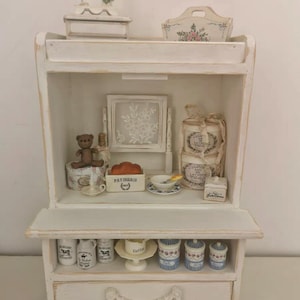 May include: A white, miniature display cabinet with multiple shelves, filled with small decorative items. Includes a sewing machine, a teddy bear, and various jars and containers. The cabinet has a distressed, vintage aesthetic.