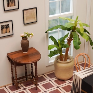 May include: A brown wooden table with a rounded top and turned legs sits in a room with white walls and a tiled floor. A vase with pink flowers sits on the table. A potted banana plant sits in the corner of the room.