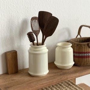 May include: A collection of miniature kitchen utensils, including wooden spatulas and a honey dipper, are displayed in a cream-colored ceramic jar. A small, cream-colored jar and a woven basket are also visible on a wooden shelf.