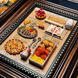 Könnte beinhalten: Ein gerahmtes Miniatur-Food-Diorama mit einer Vielzahl von Gerichten. Enthält eine Erdbeertorte, Pizza, einen Teller mit Essen, ein Kochbuch und eine Gemüsekonserve. Der Rahmen ist dunkelbraun mit goldenen Akzenten.