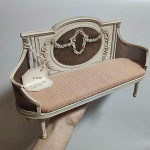 Puede incluir: Un banco en miniatura de estilo antiguo con marco color crema, asiento rosa y respaldo de malla marrón. El banco presenta tallas florales ornamentadas y una etiqueta que dice "T ONEX".