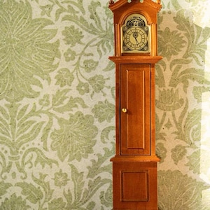 Puede incluir: Un reloj de pie de madera con una esfera dorada y marcadores de hora romanos negros. El reloj tiene una caja de madera marrón con una sola puerta y una base con un diseño decorativo. El reloj está de pie sobre un suelo de madera frente a un papel tapiz floral verde.