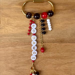 May include: A gold-toned oval carabiner keychain featuring a variety of dangling charms. The charms include white letter beads spelling GO DAWGS, interspersed with small red, black, and white beads, a gold metal chain, and a cluster of beads at the base.