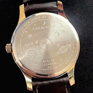 May include: A gold-toned watch with a brown leather strap. The watch face features the text "DESIGNED BY BULOVA CARAVELLE" and a solar system design. The back of the watch is stainless steel and water resistant.