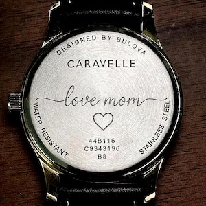 May include: A stainless steel Caravelle watch back with a black leather strap. The watch face is engraved with "love mom" and a heart symbol. The watch is water resistant and designed by Bulova.