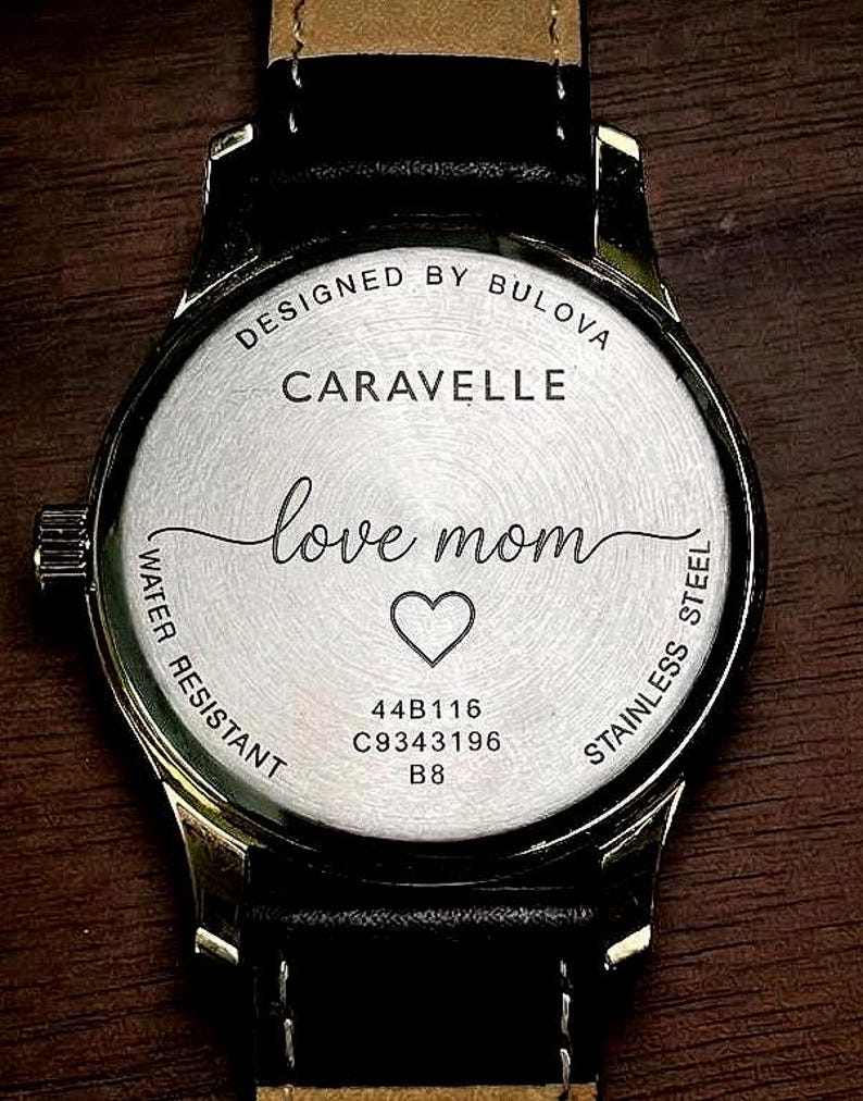 May include: A stainless steel Caravelle watch back with a black leather strap. The watch face is engraved with "love mom" and a heart symbol. The watch is water resistant and designed by Bulova.