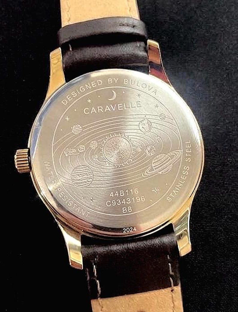 May include: A gold-toned watch with a brown leather strap. The watch face features the text "DESIGNED BY BULOVA CARAVELLE" and a solar system design. The back of the watch is stainless steel and water resistant.