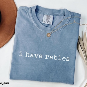 May include: A blue jean Comfort Colors t-shirt with the text "i have rabies" in white. A gold necklace with a pendant is on the shirt. A brown hat is in the upper left corner.
