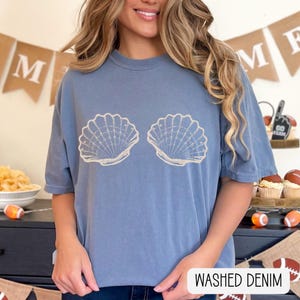 May include: A washed denim blue short-sleeve t-shirt featuring a graphic print of two beige seashells centered on the chest. The shirt has a crew neckline and a relaxed fit. The text "WASHED DENIM" is printed in black in the bottom right corner.